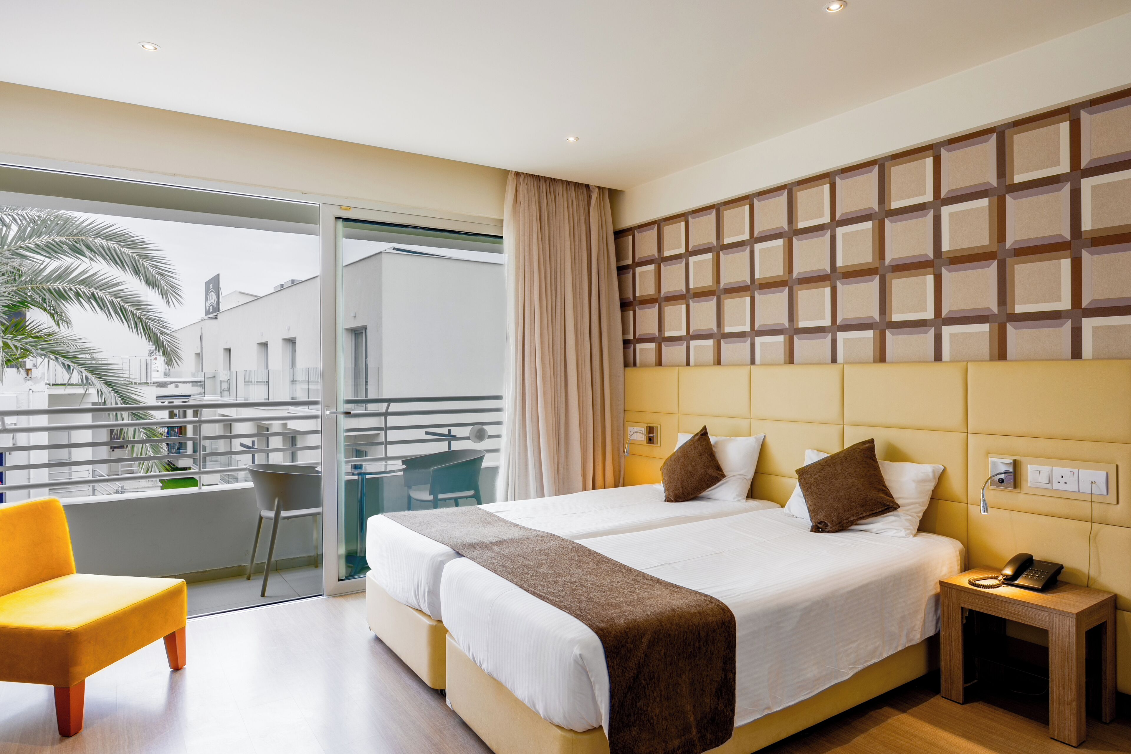 A hotel room with a large bed, a small bedside table with a phone, a chair, and a balcony with a view of the city.