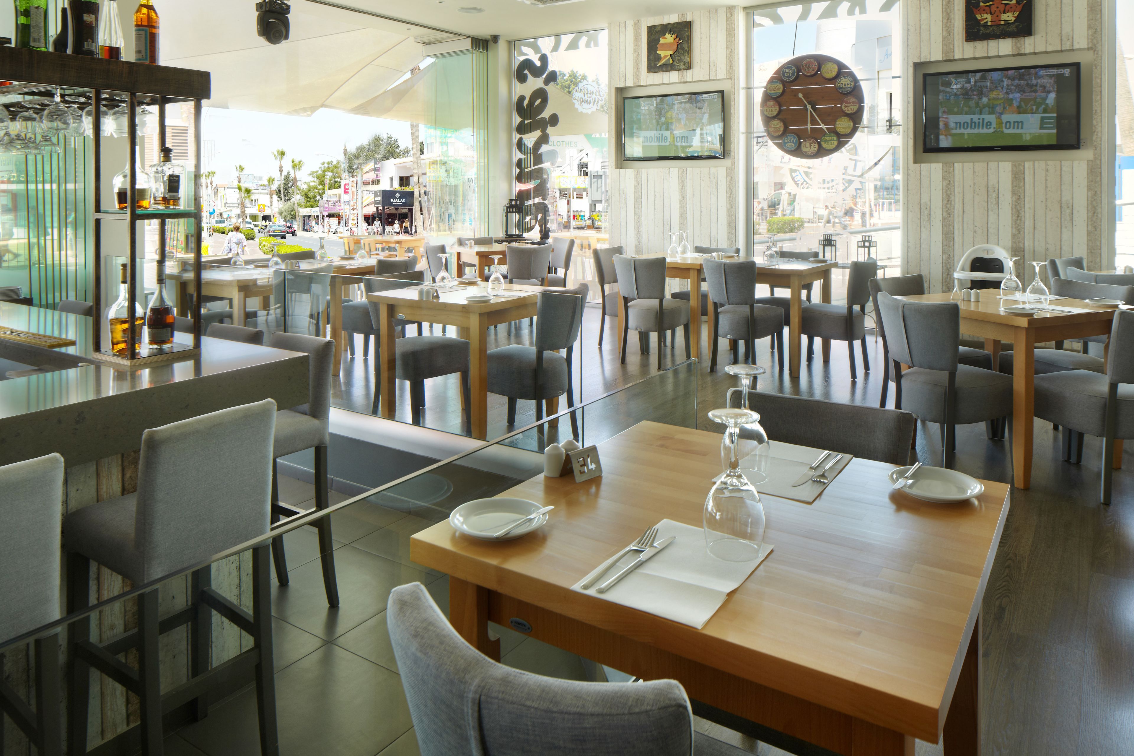 A restaurant with multiple tables and chairs, a bar area with bottles, and a large clock on the wall.