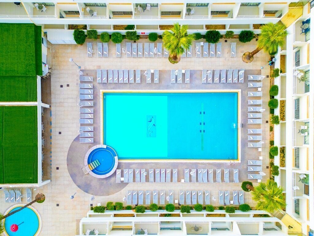 Vue aérienne d’un hôtel doté d’une piscine centrale, de plusieurs espaces de détente et d’une pelouse verdoyante.