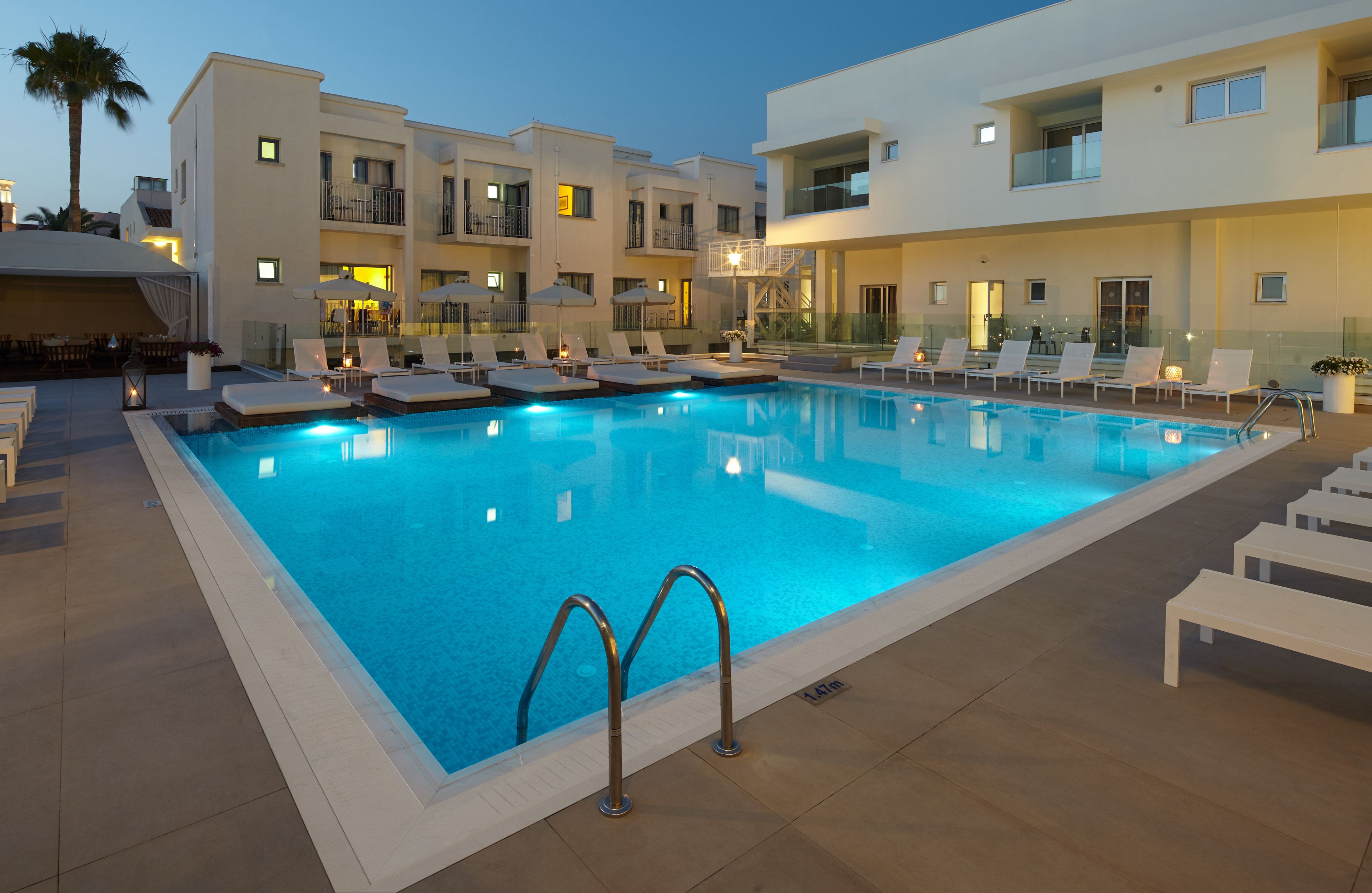 Piscina con sillas de descanso y sombrillas, rodeada de edificios modernos con balcones e iluminación.