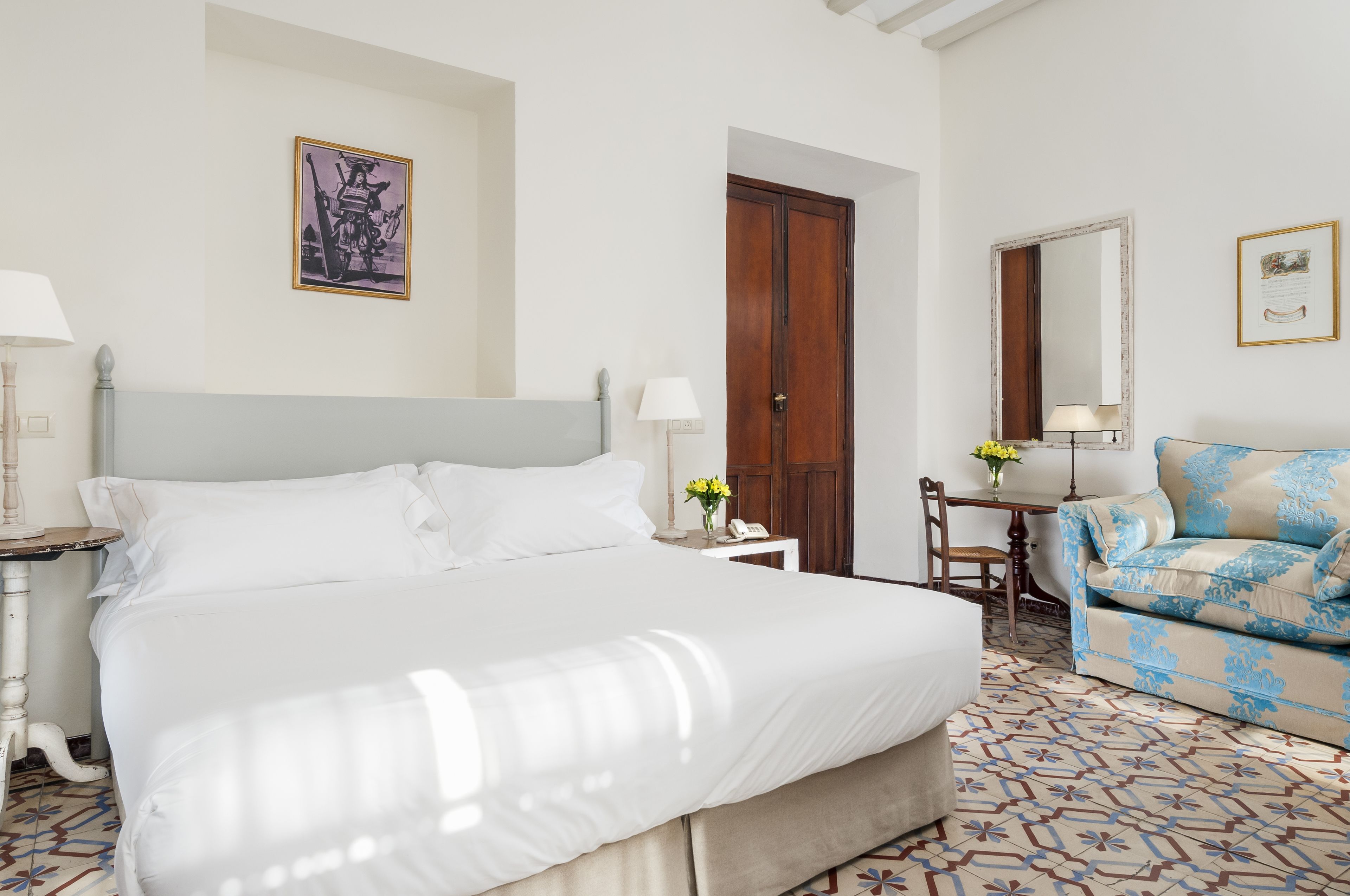 Een hotelkamer met een groot bed, een klein tafeltje met een lamp, een stoel, een bank en een deur.
