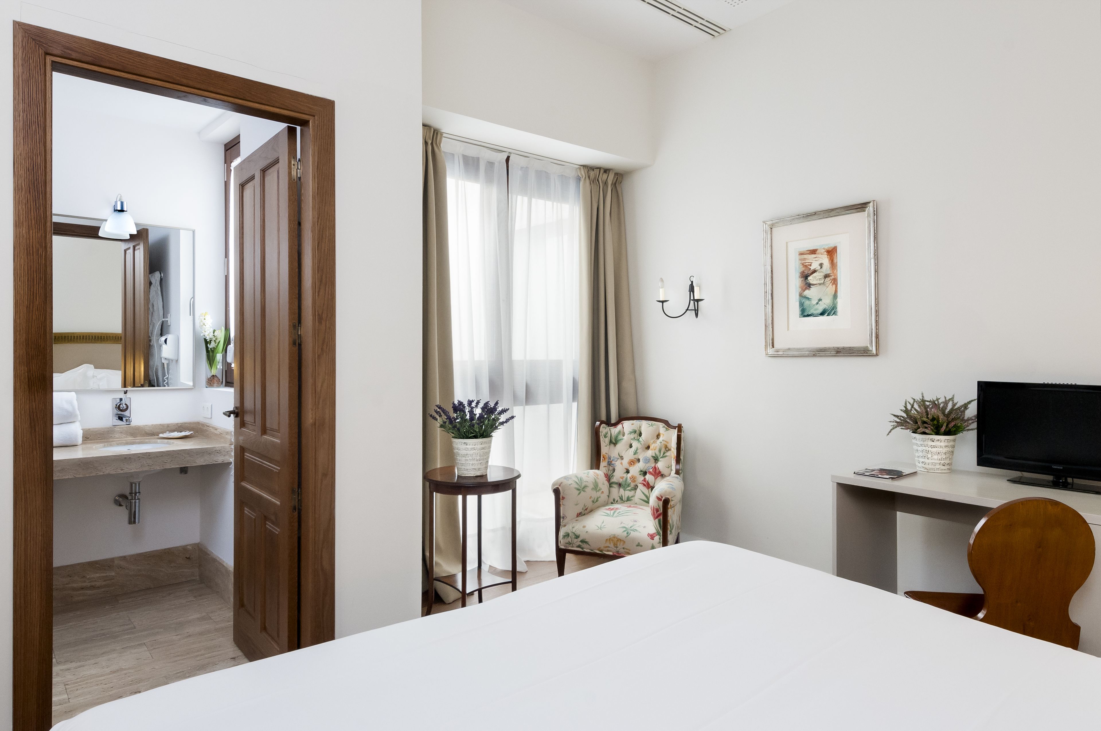 A hotel room with a bed, a desk with a computer, a chair, a small table with plants, a mirror, and a door leading to a bathroom.