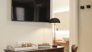 Una habitación con un escritorio de madera, una silla, un televisor montado en la pared y un espejo que refleja un baño con un lavamanos y un perchero.