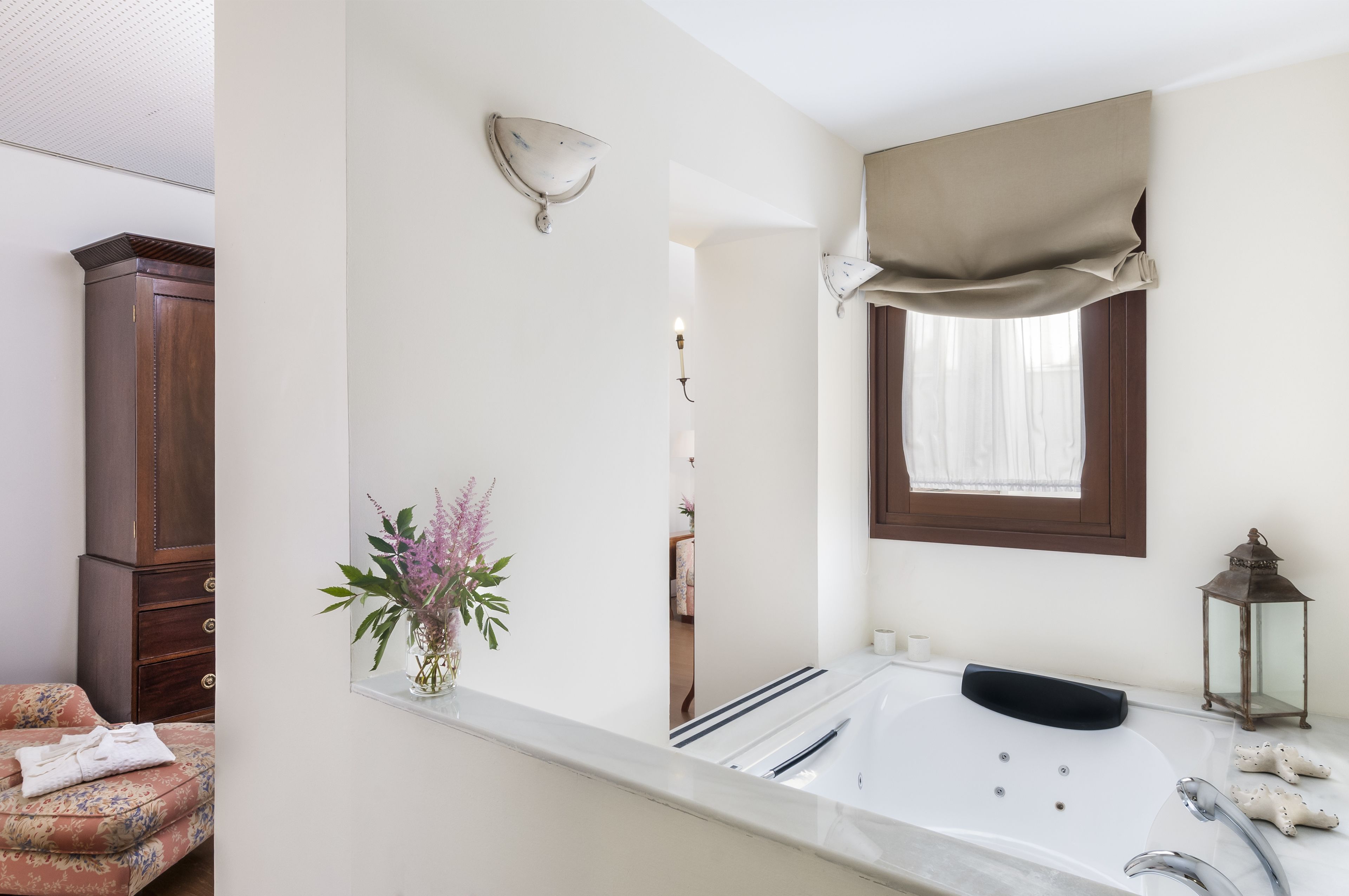 A bathroom with a bathtub, a mirror, a window with a beige curtain, a wooden cabinet, and a vase with flowers.