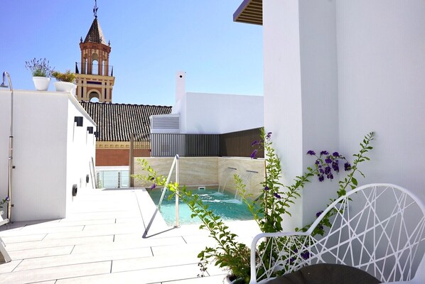 A rooftop pool area with a view of a bell tower, surrounded by white walls and potted plants.