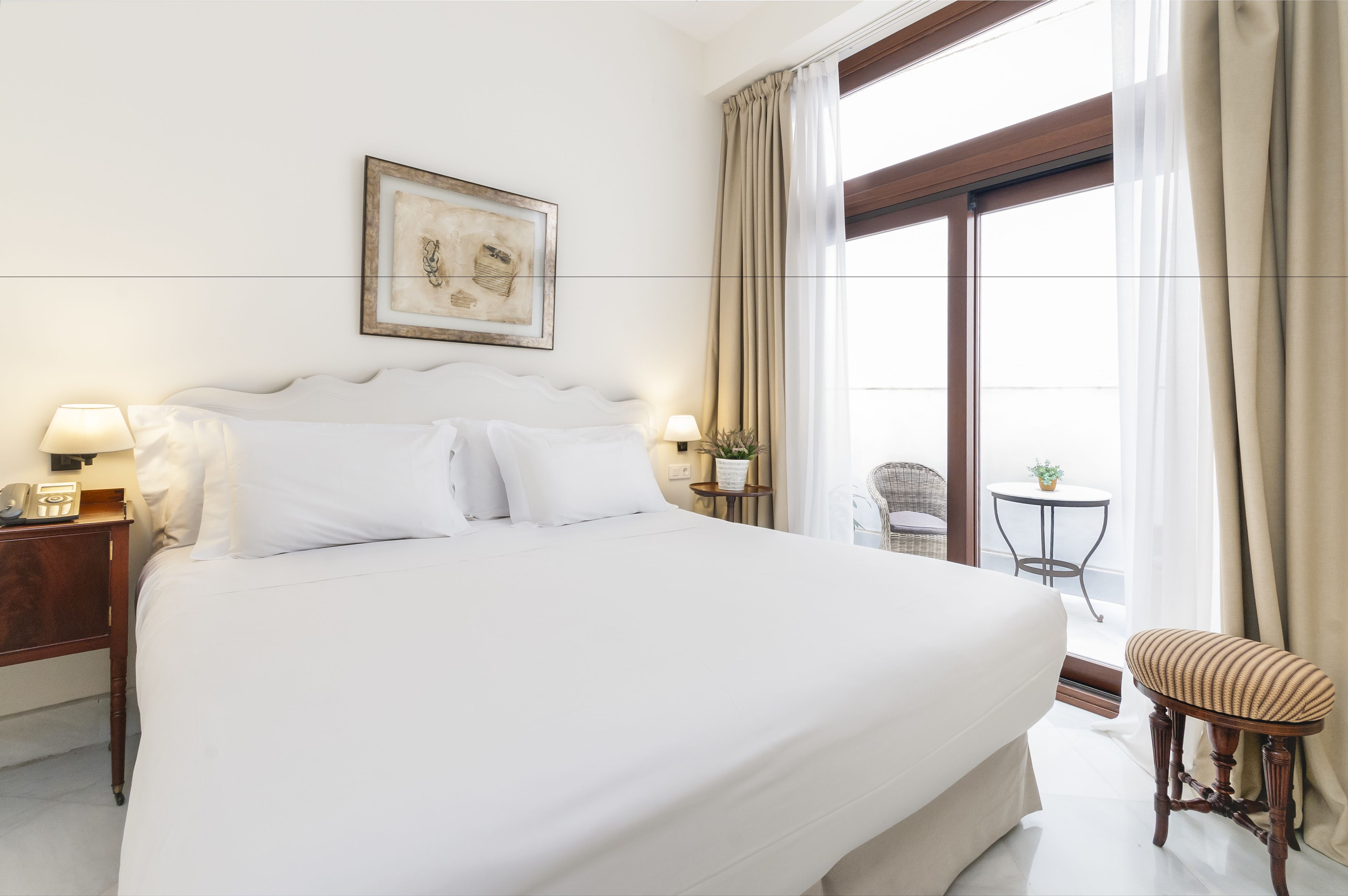 A large bed with white linens, a bedside table with a lamp, a framed picture on the wall, and a view of a balcony with a chair and a small table.