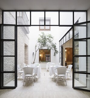An open-plan dining area with white wicker chairs and tables, a brick wall, and a view of the street through large windows.