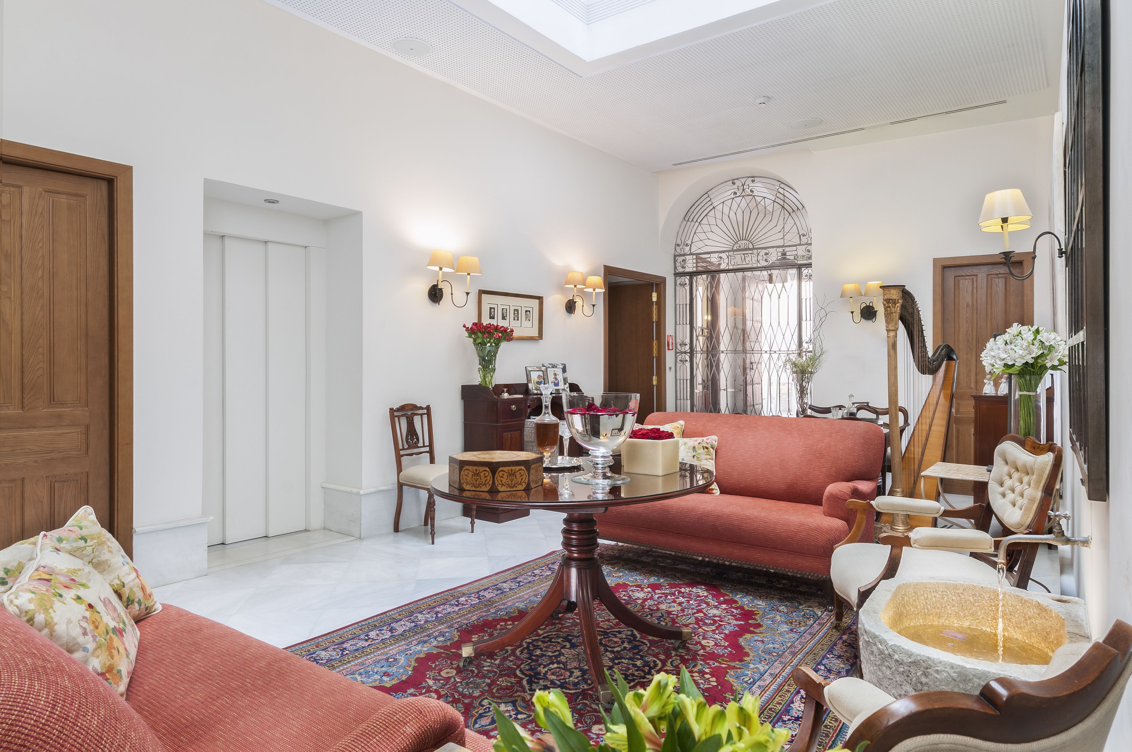 A spacious living room with a large red sofa, a glass coffee table, and a patterned rug. There is a wooden door, a white wall, and a decorative mirror.