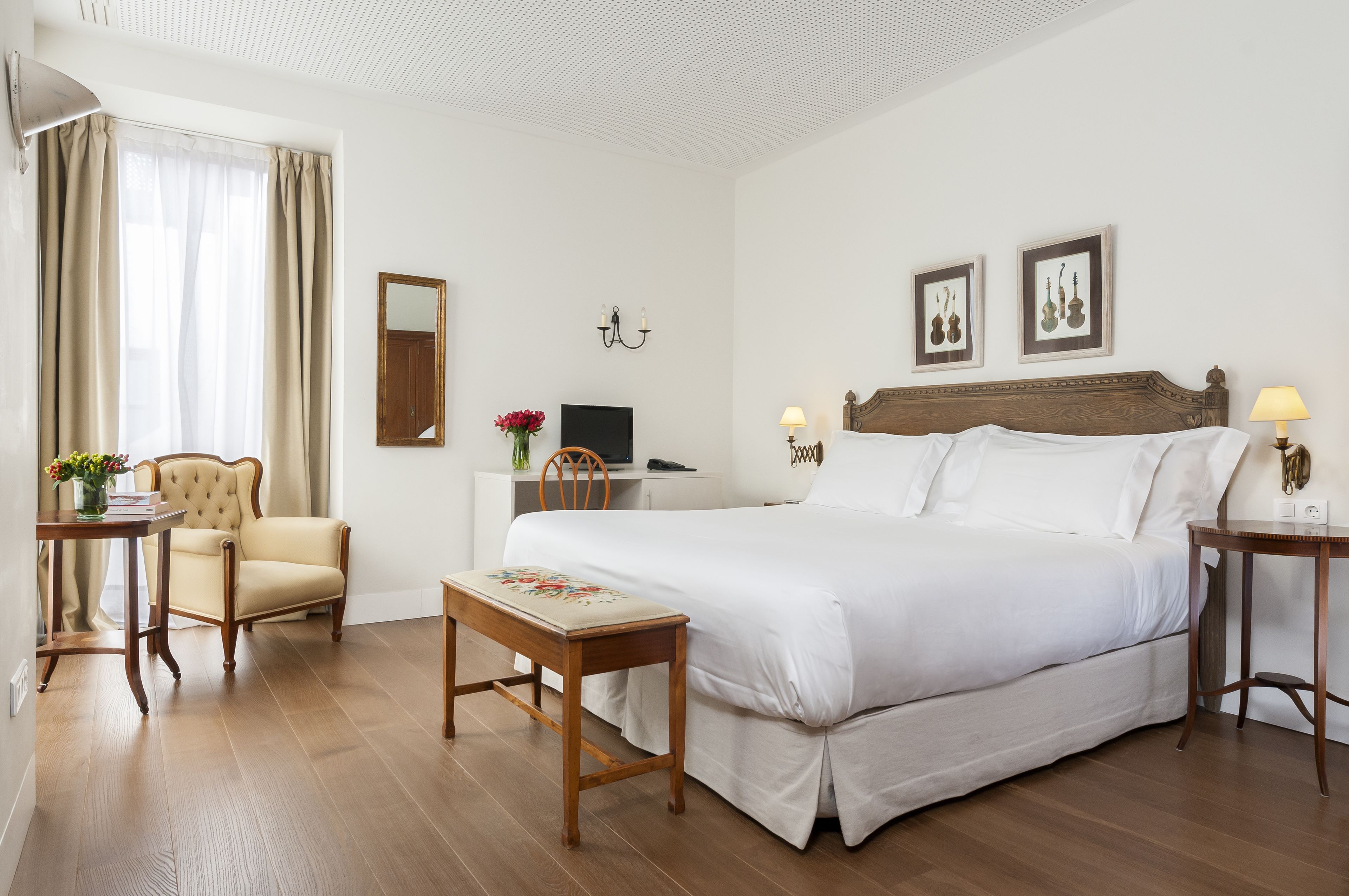 Habitación de hotel con una cama grande, una silla de madera, una mesita pequeña, un escritorio, un espejo y dos lámparas de pared.