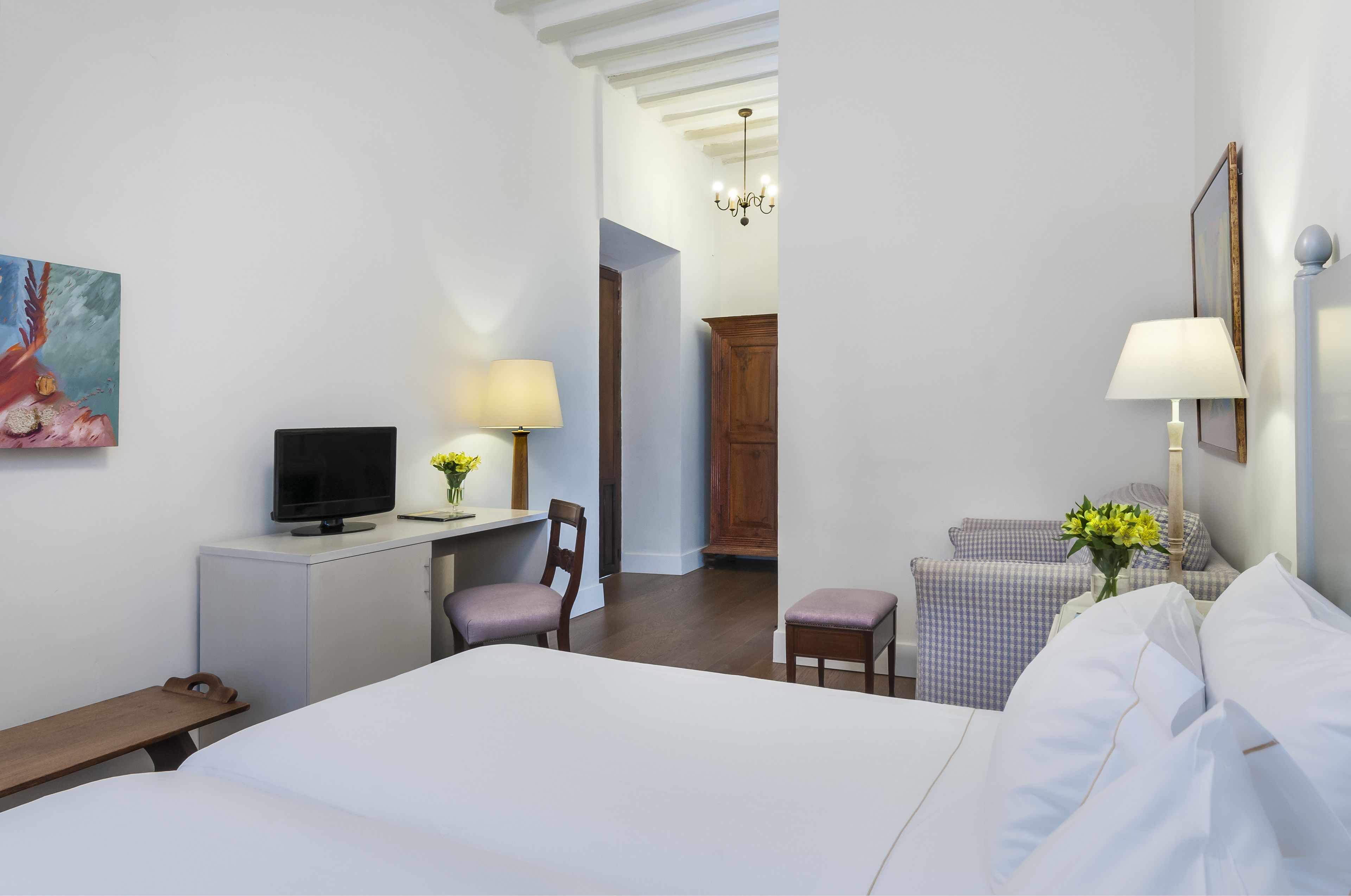 A hotel room with a bed, a desk with a TV, a chair, a small table, a lamp, and a painting on the wall.