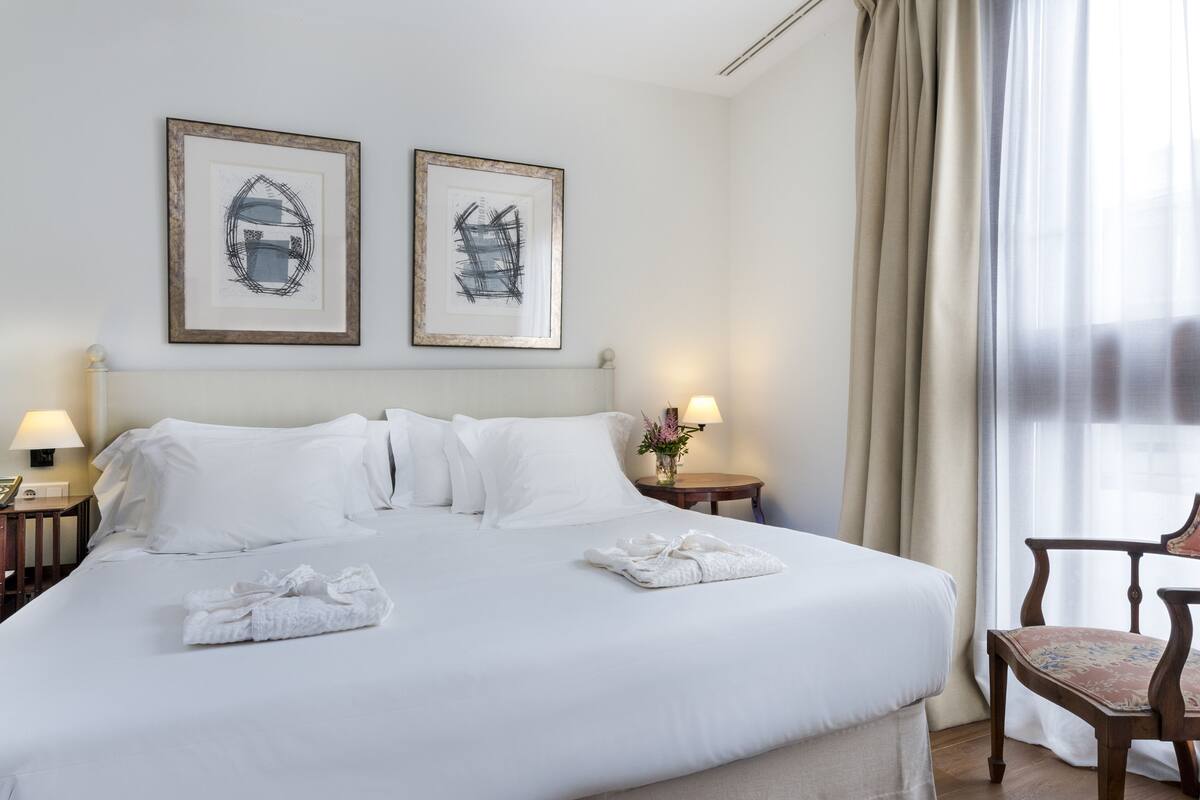Une chambre d’hôtel avec un grand lit, deux œuvres d’art encadrées au mur, une table de chevet avec une lampe et une chaise près de la fenêtre.