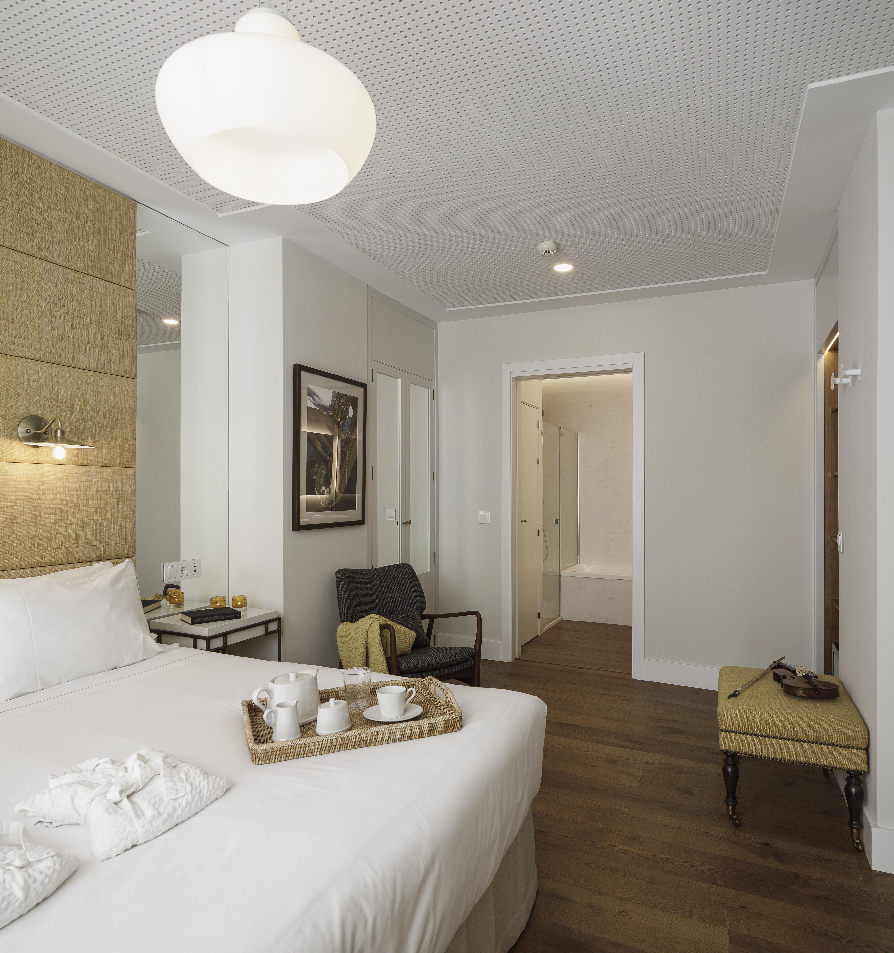 A hotel room with a large bed, a bedside table with a lamp, a chair, a small table with a tray of items, and a mirror.