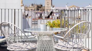 A rooftop terrace with white wire mesh chairs and a small table, overlooking a cityscape with historic buildings and a tower.