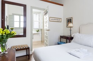 A hotel room with a bed, a bedside table with a lamp, a small stool, a mirror, and a door leading to a bathroom.