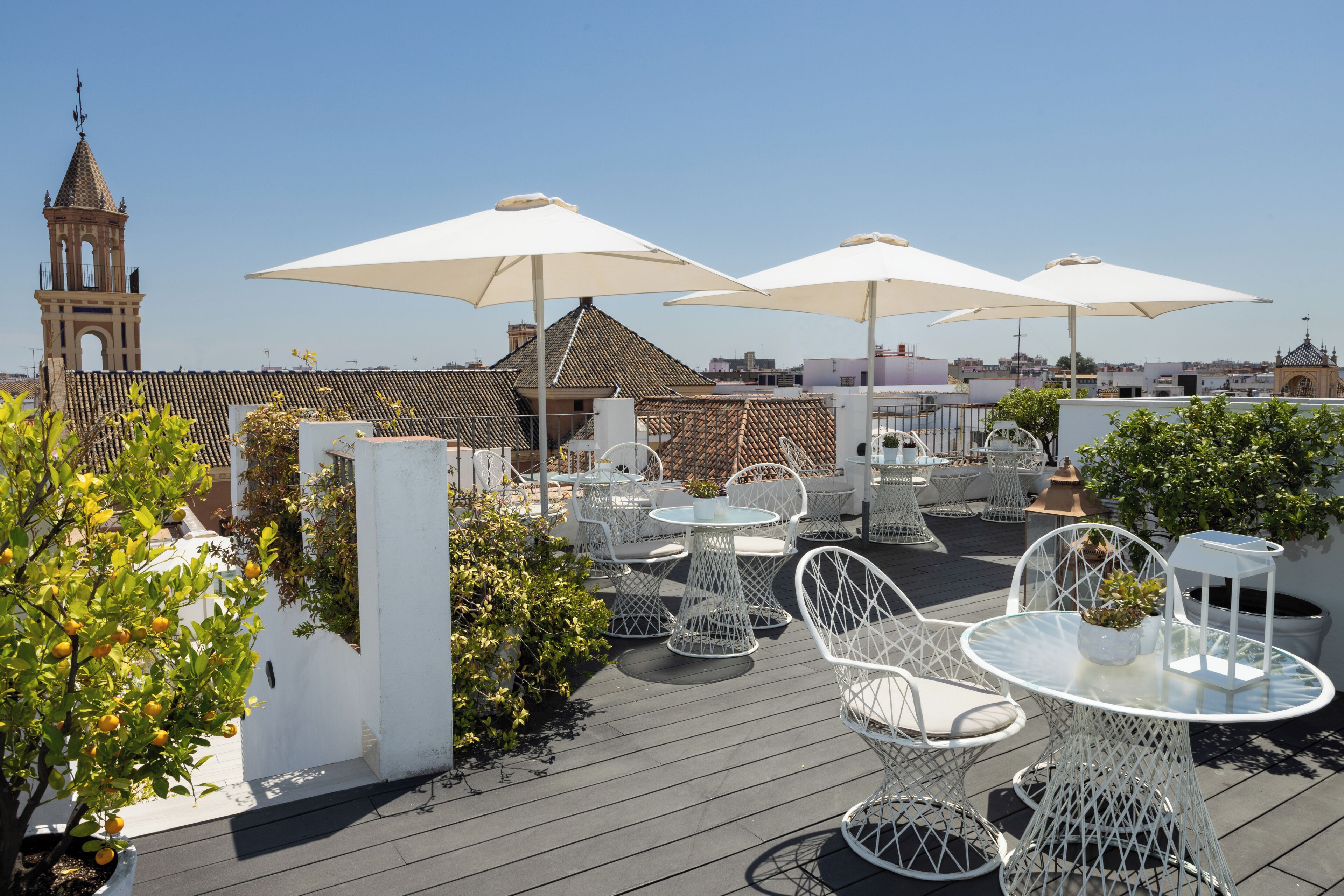 A rooftop terrace with white metal chairs and tables, large umbrellas, and a view of a church tower.