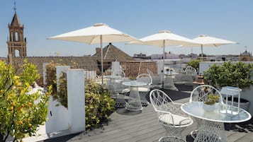 A rooftop terrace with white metal chairs and tables, large umbrellas, and a view of a church tower.