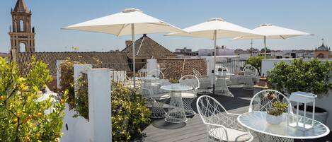 A rooftop terrace with white metal chairs and tables, large umbrellas, and a view of a church tower.