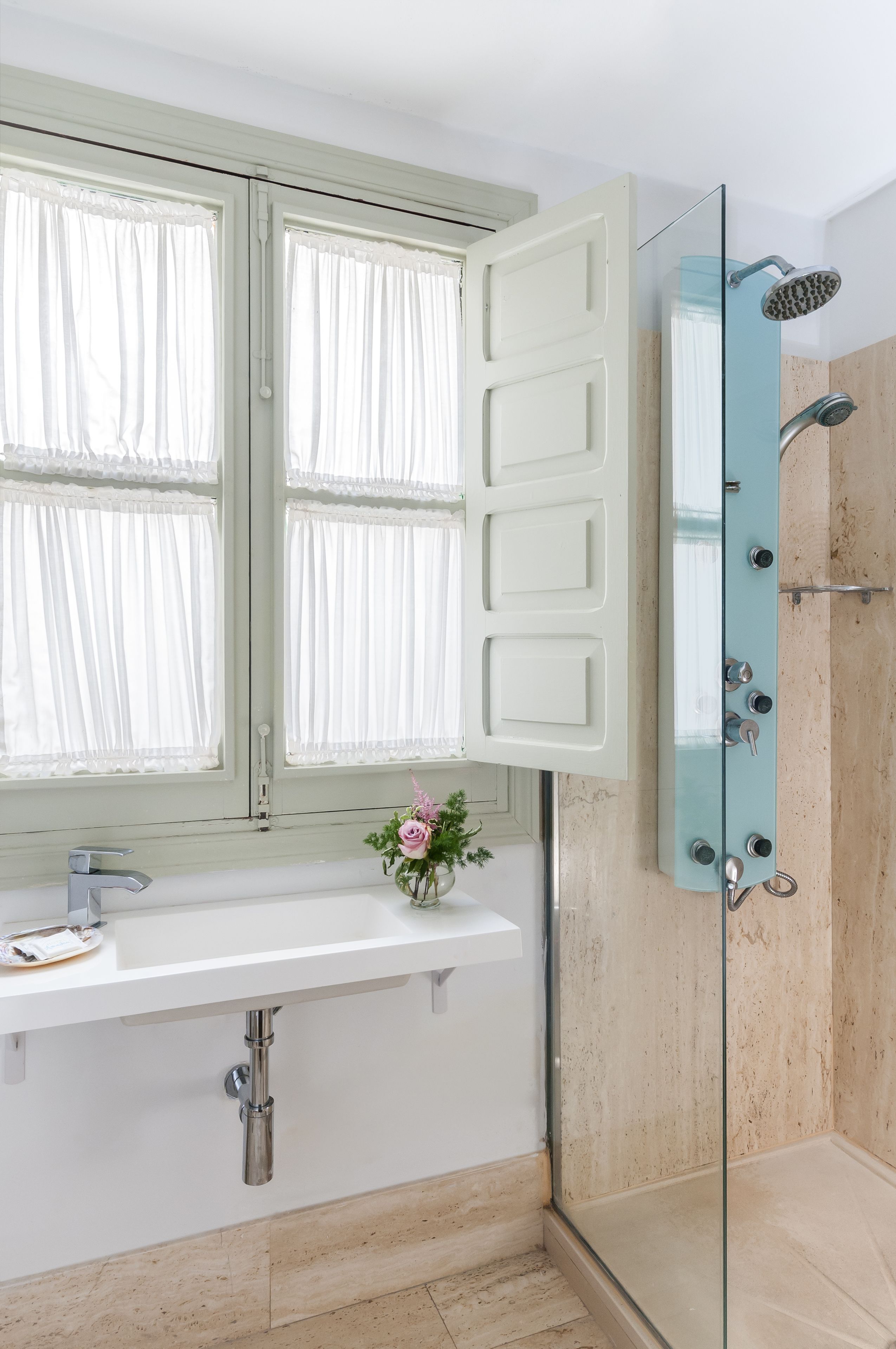 Une salle de bain avec un lavabo blanc, une cabine de douche en verre et une fenêtre agrémentée de rideaux blancs.