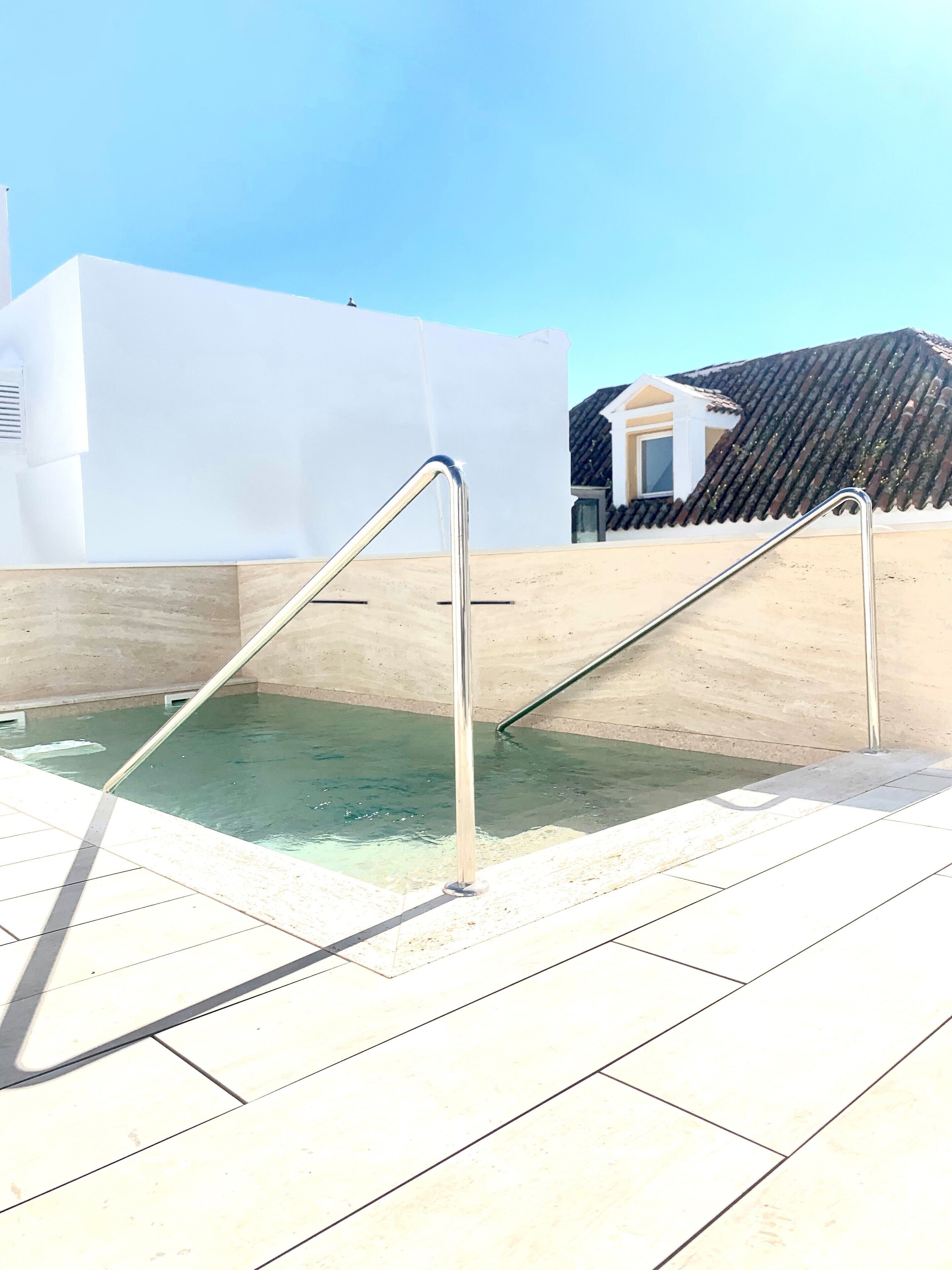 A swimming pool with metal railings, a tiled deck, and a building with a tiled roof in the background.
