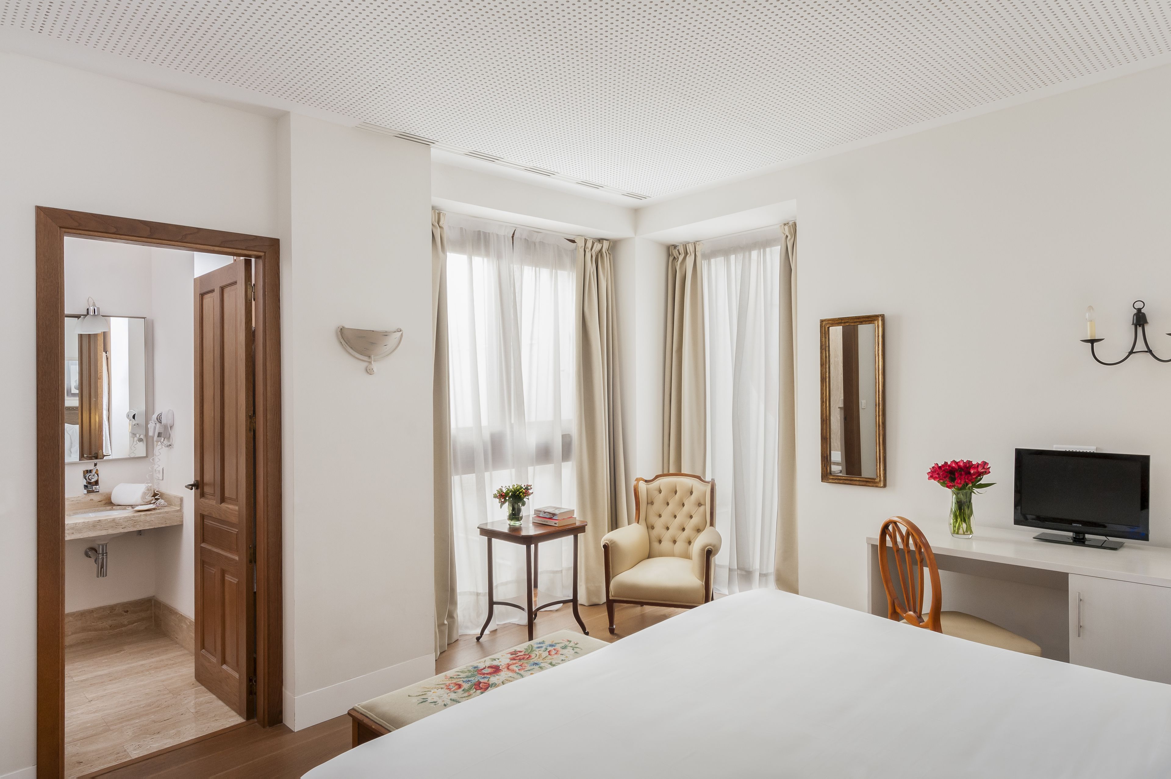 Habitación de hotel con una cama, un escritorio con televisor, una silla, una mesita con un jarrón de flores y un baño visible a través de una puerta abierta.