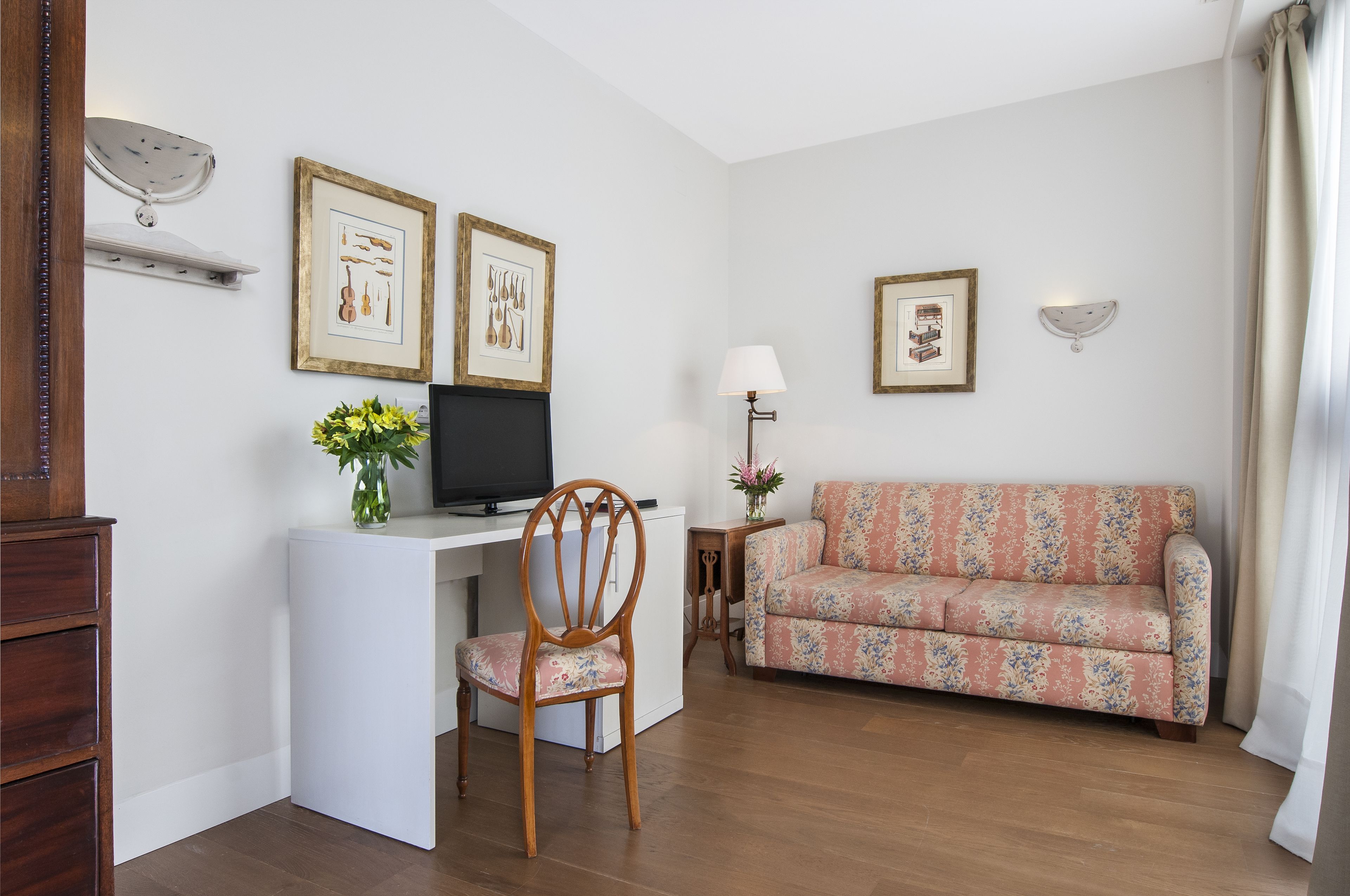 A living room with a wooden desk, a television, a floral sofa, and framed pictures on the wall.