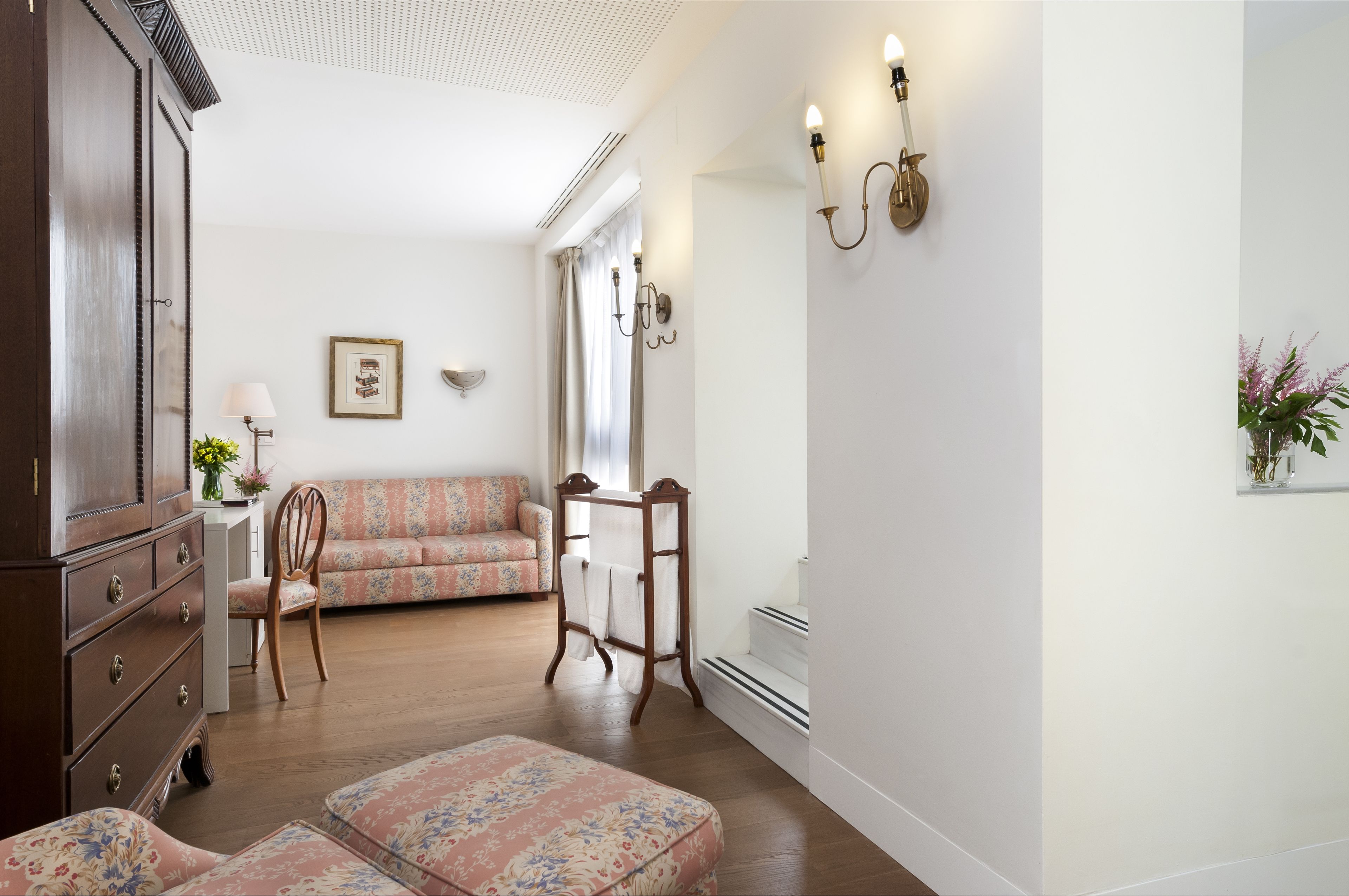A room with a wooden dresser, a floral sofa, a wooden chair, a small table with a lamp, and a wall-mounted light fixture.