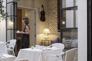 Un espacio de comedor al aire libre con mesas blancas y sillas de mimbre, un violín en la pared y una lámpara sobre una pequeña mesa.