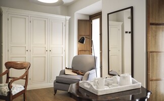 Une chambre d’hôtel avec une chaise en bois, un fauteuil gris, un bureau avec un plateau contenant des articles de toilette et un grand miroir.