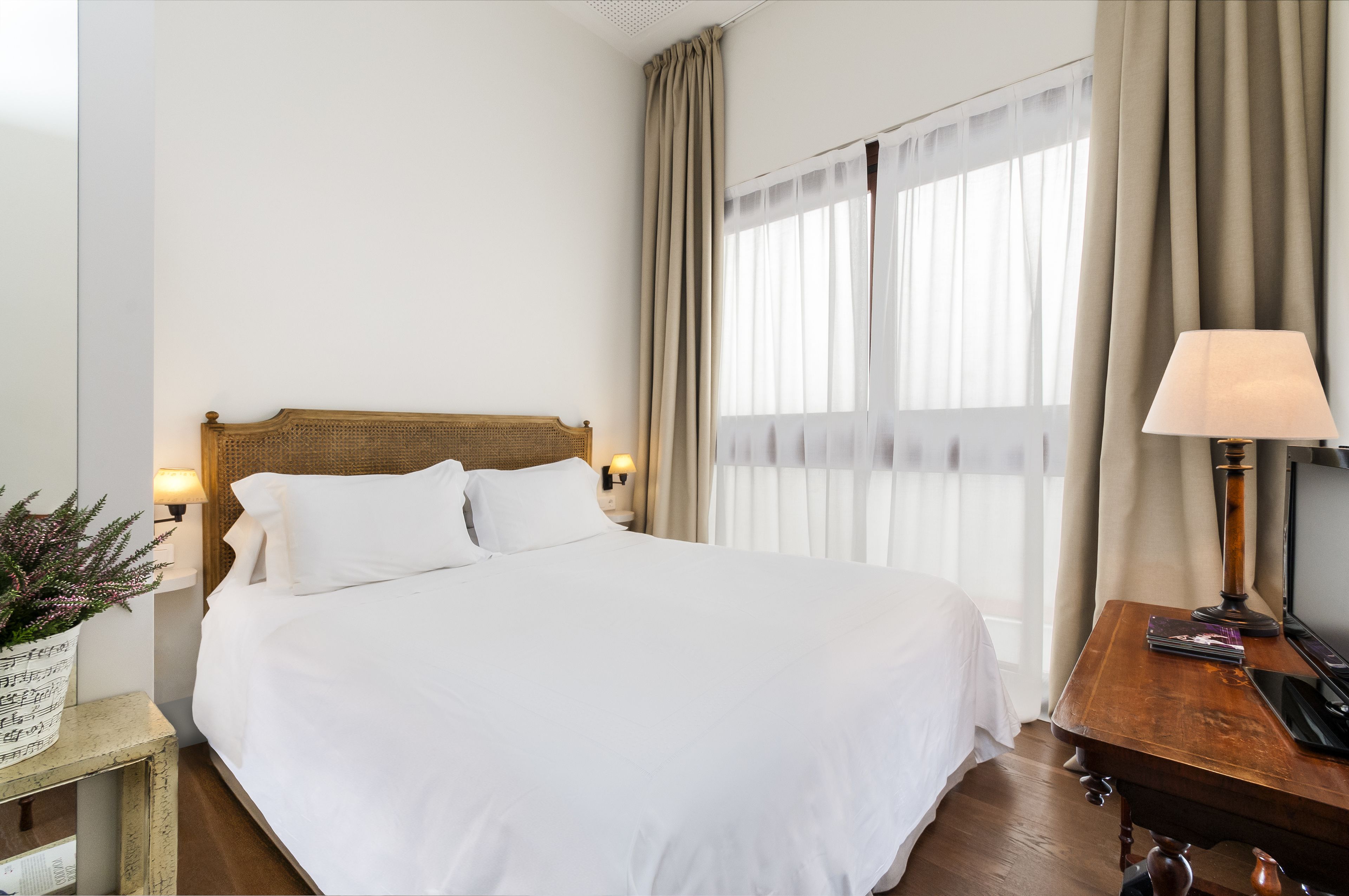 A neatly made bed with white linens, a wooden headboard, a bedside table with a lamp, and a window with sheer curtains.