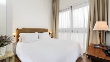 A neatly made bed with white linens, a wooden headboard, a bedside table with a lamp, and a window with sheer curtains.