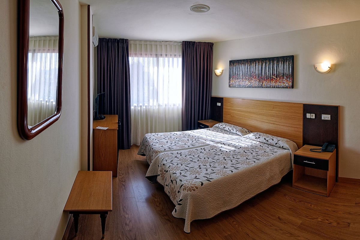 A hotel room with a wooden bed, a desk, a mirror, a painting, and a nightstand with a phone.