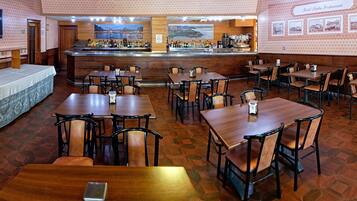 Un restaurante con varias mesas y sillas, una zona de bar con mostrador y diferentes botellas, y un televisor montado en la pared.
