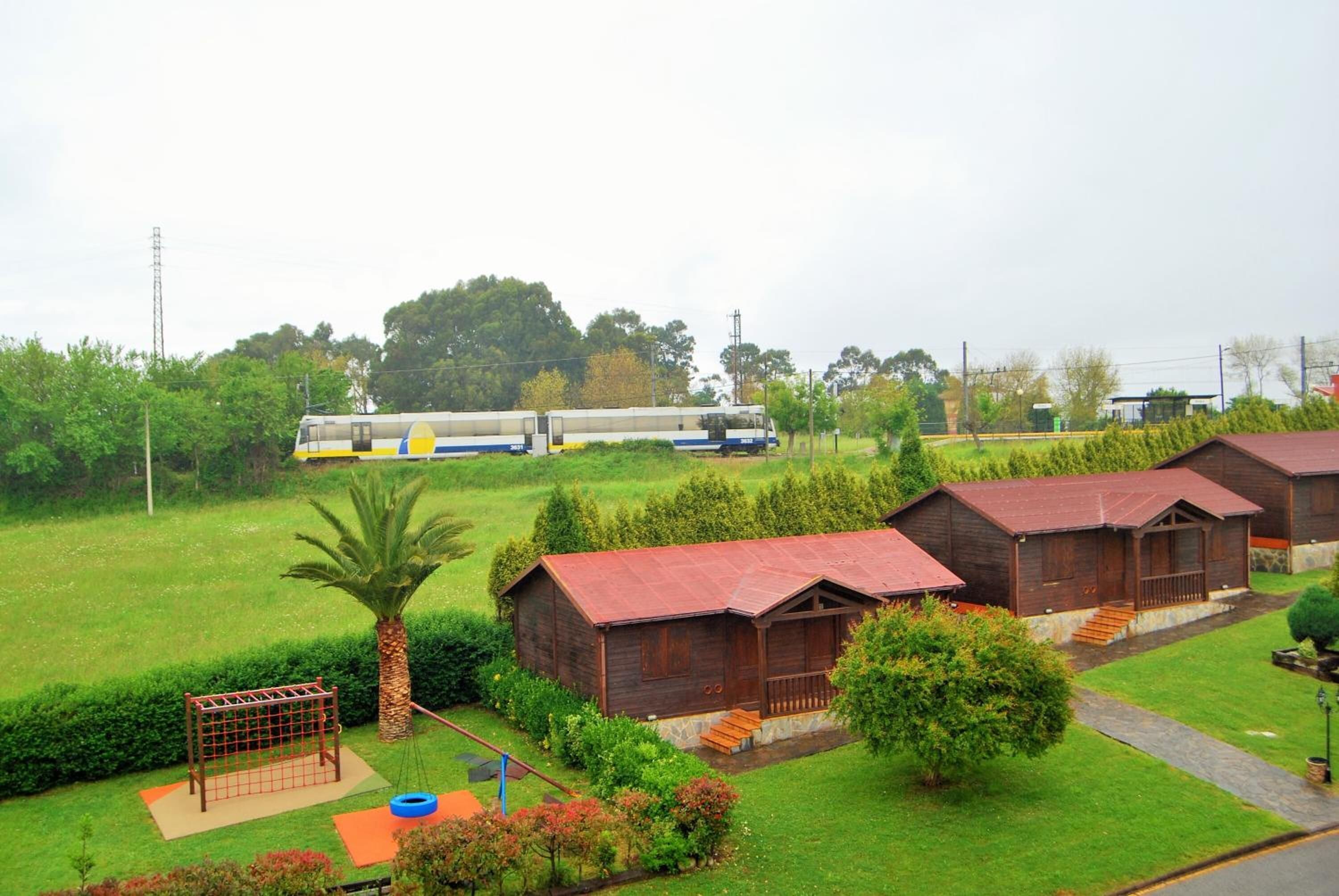 Un entorno rural con cabañas de madera, un parque infantil y un tren al fondo.