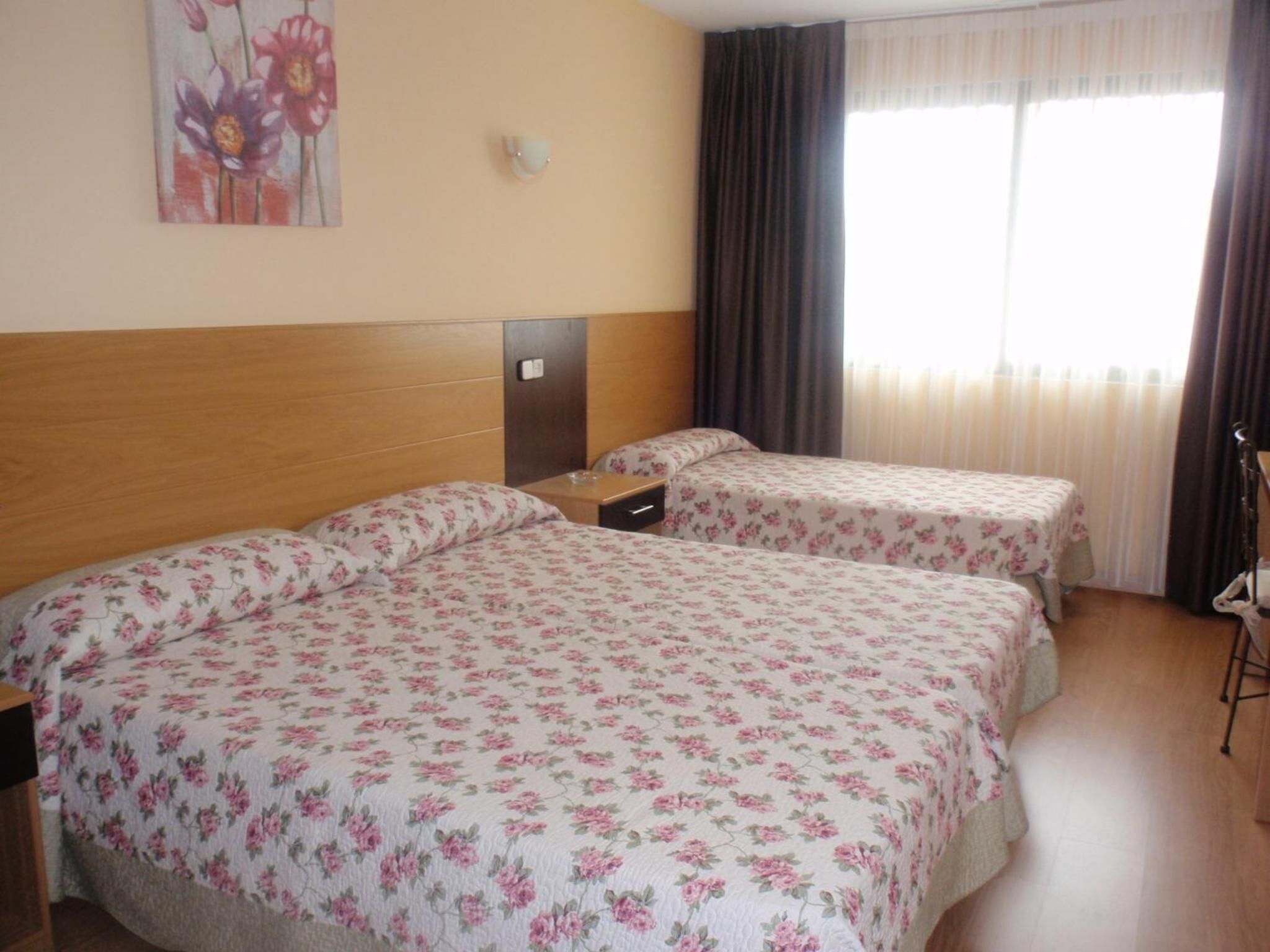 Habitación de hotel con dos camas, cabecera de madera, una mesita, un buró, un cuadro en la pared y una ventana con cortinas.
