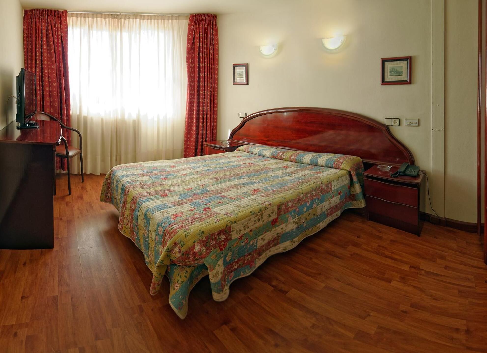 A bedroom with a wooden bed, a desk, and a chair. There are curtains on the window, and a framed picture on the wall.
