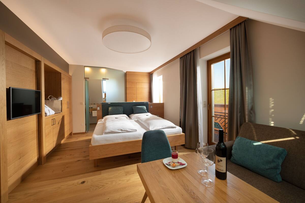 Une chambre d’hôtel moderne avec un plancher en bois, un lit avec deux oreillers, un canapé avec un coussin, une table avec une bouteille et des verres, et un téléviseur fixé au mur.