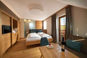 Una habitación de hotel moderna con piso de madera, una cama con dos almohadas, un sofá con un cojín, una mesa con una botella y vasos, y un televisor en la pared.