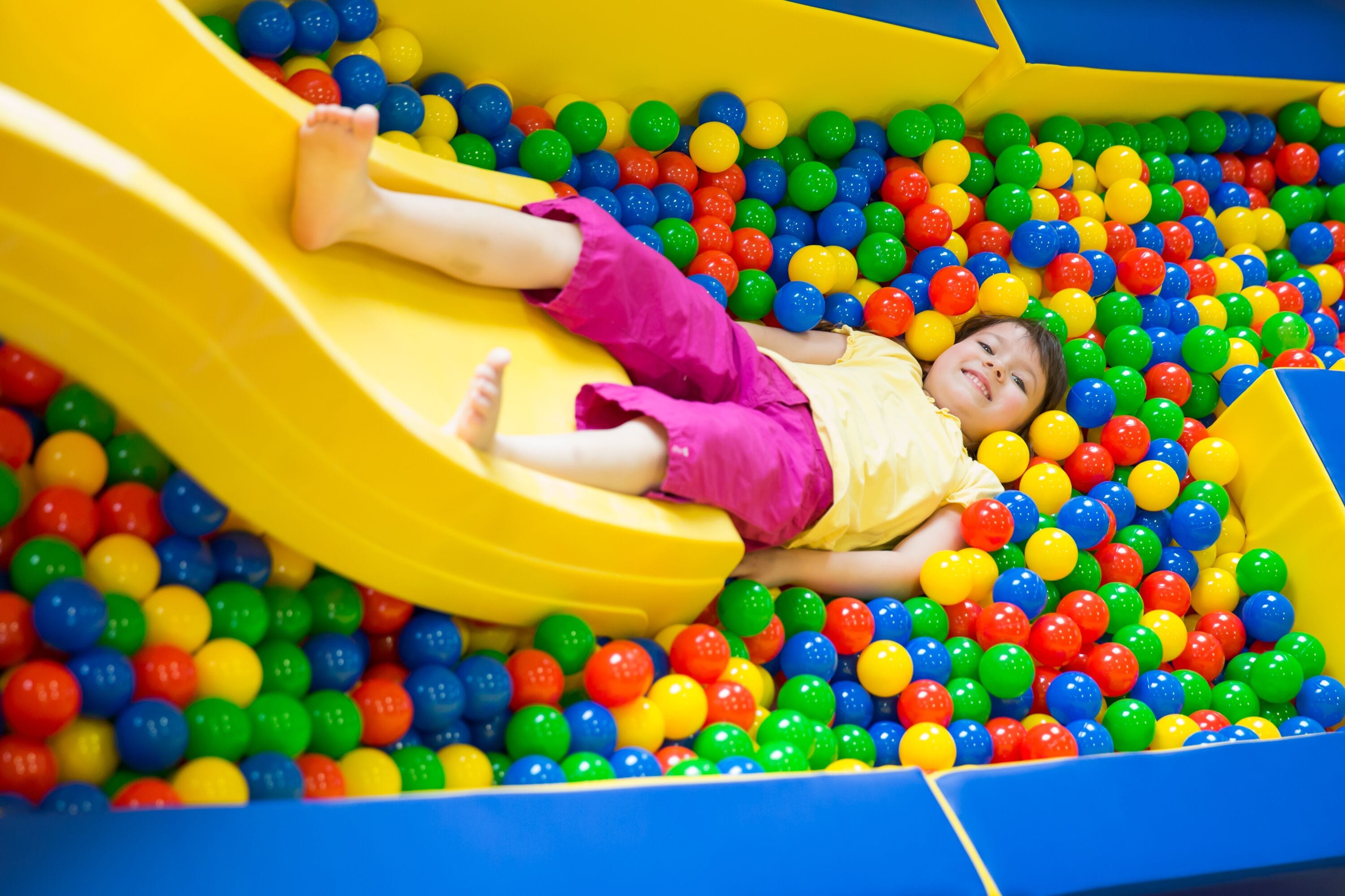 A child lying in a ball pit with a yellow slide.