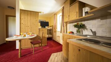 A compact kitchen with wooden cabinets, a dining area with a red carpet, and a television in the background.