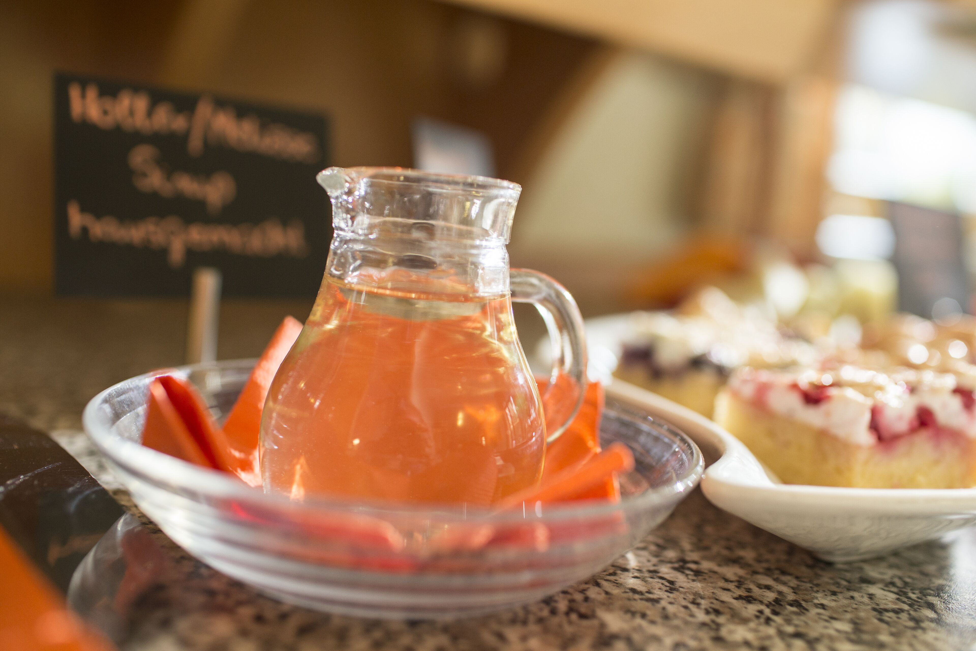 A clear glass pitcher with a straw, filled with a red beverage, placed on a plate with carrot sticks. In the background, there is a sign with text and a dessert on a plate.