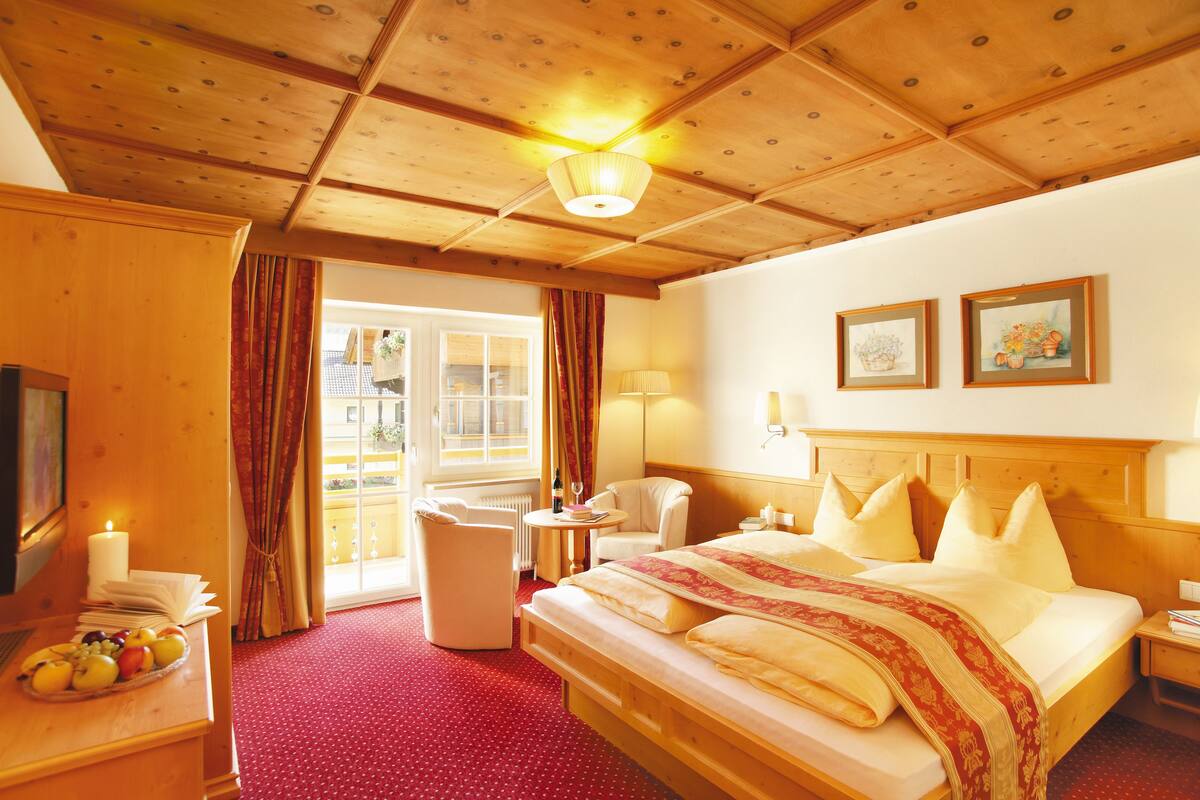 A hotel room with a wooden ceiling, a large bed with pillows, a bedside table, a chair, a small table with a lamp, and a window with curtains.