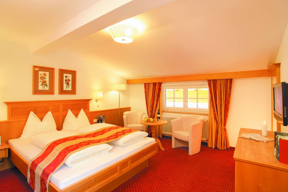 A hotel room with a wooden bed, a desk, a chair, a small table with a lamp, and a television.