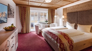 A hotel room with a wooden ceiling, a bed with a red and gold bedspread, a wooden dresser, a desk with a lamp, and a view of the mountains through the window. - Hotel Alpenherz (Gerlos)