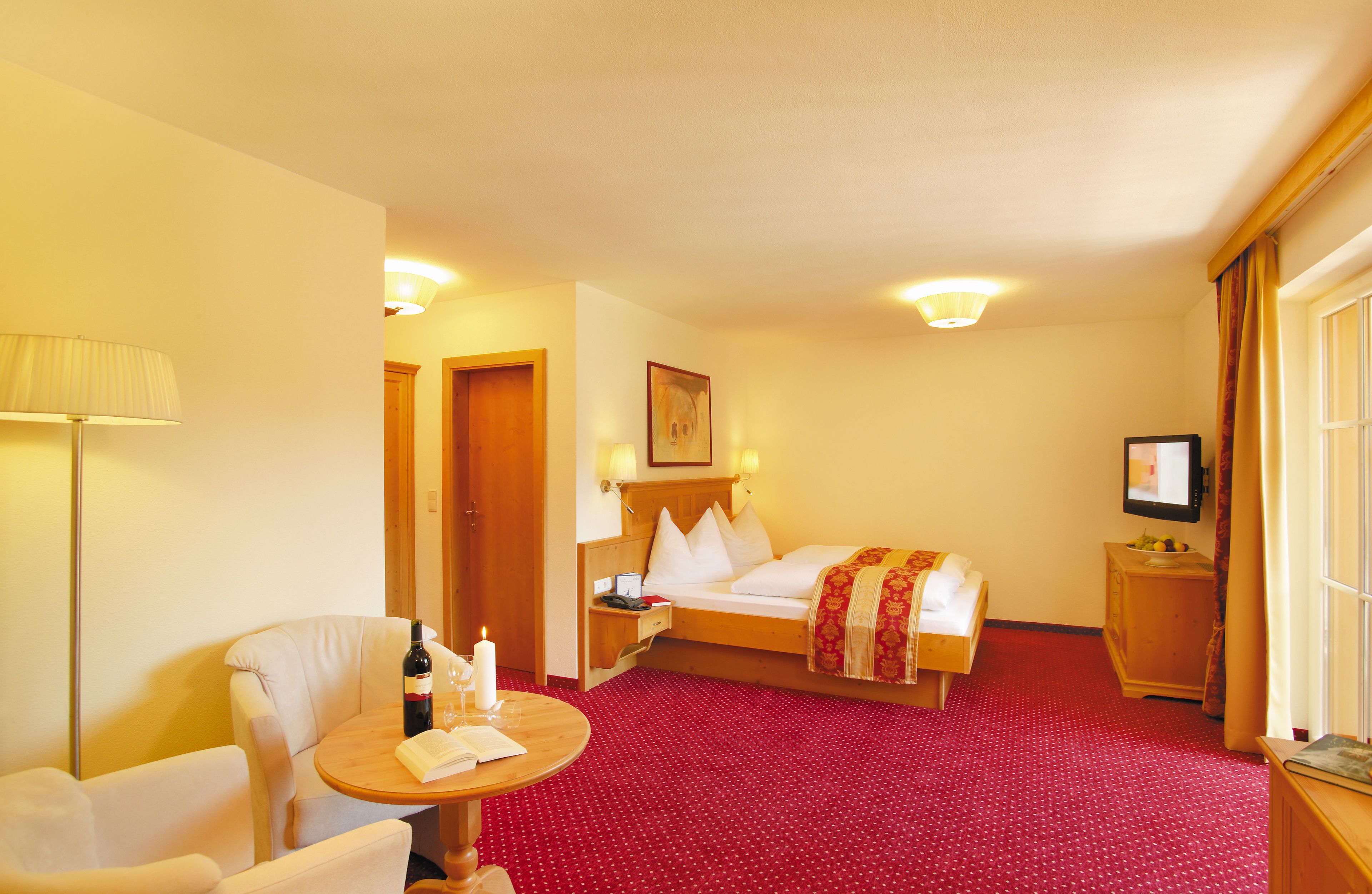 Habitación de hotel con cama, una silla, una mesita con una botella y una vela, una mesita de noche, un televisor y piso cubierto con alfombra roja.