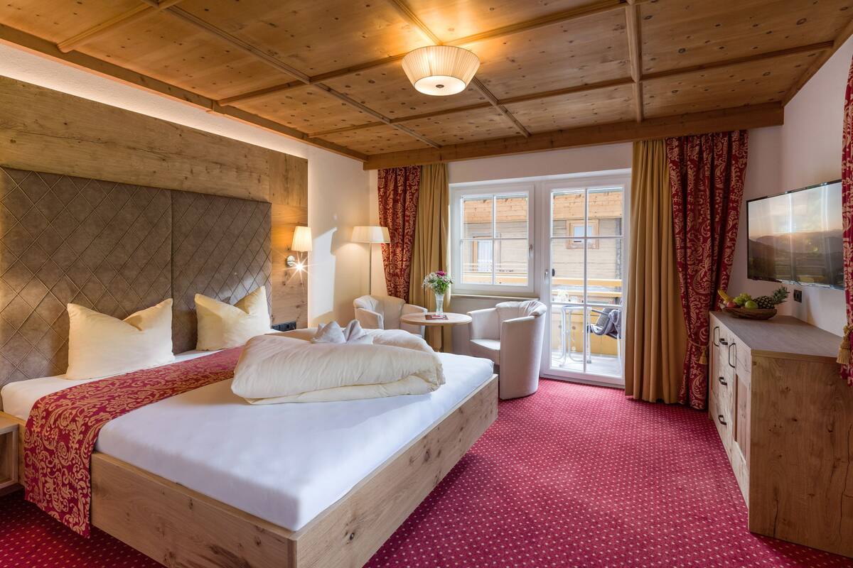 A hotel room with a wooden ceiling, a large bed with white linens, a small round table with a lamp, a TV on the wall, and a balcony with curtains.