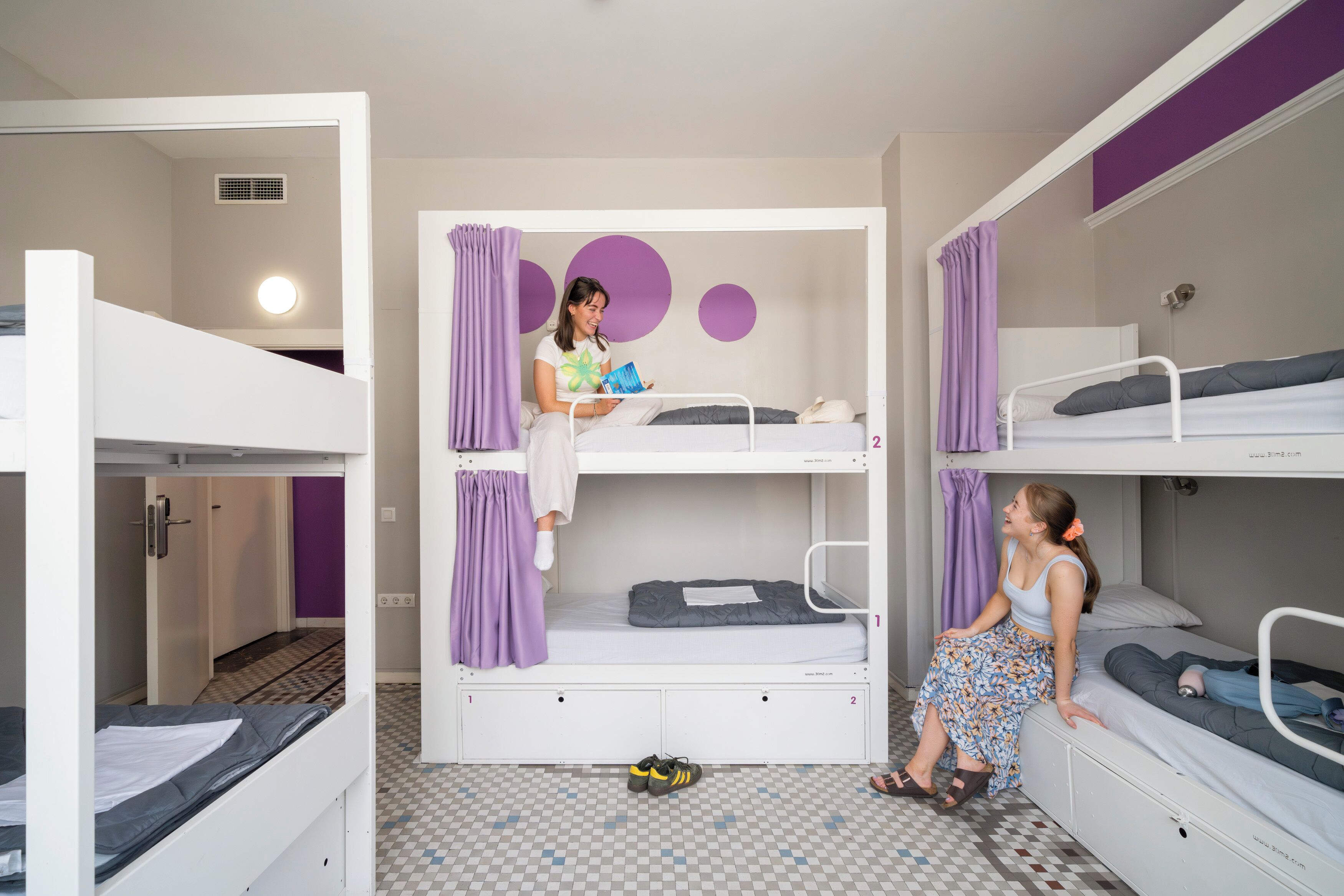Habitación con literas, una cortina morada, un ventanal pequeño y suelo a cuadros.
