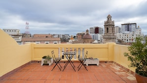 A rooftop terrace with a yellow wall, terracotta tiles, two wooden chairs, a small table, and potted plants.