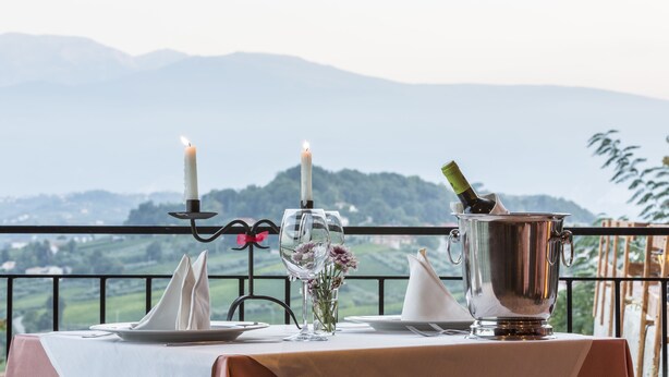 Une table dressée pour deux personnes, avec des bougies, une bouteille de vin et un pichet en argent, donnant sur un paysage montagneux.