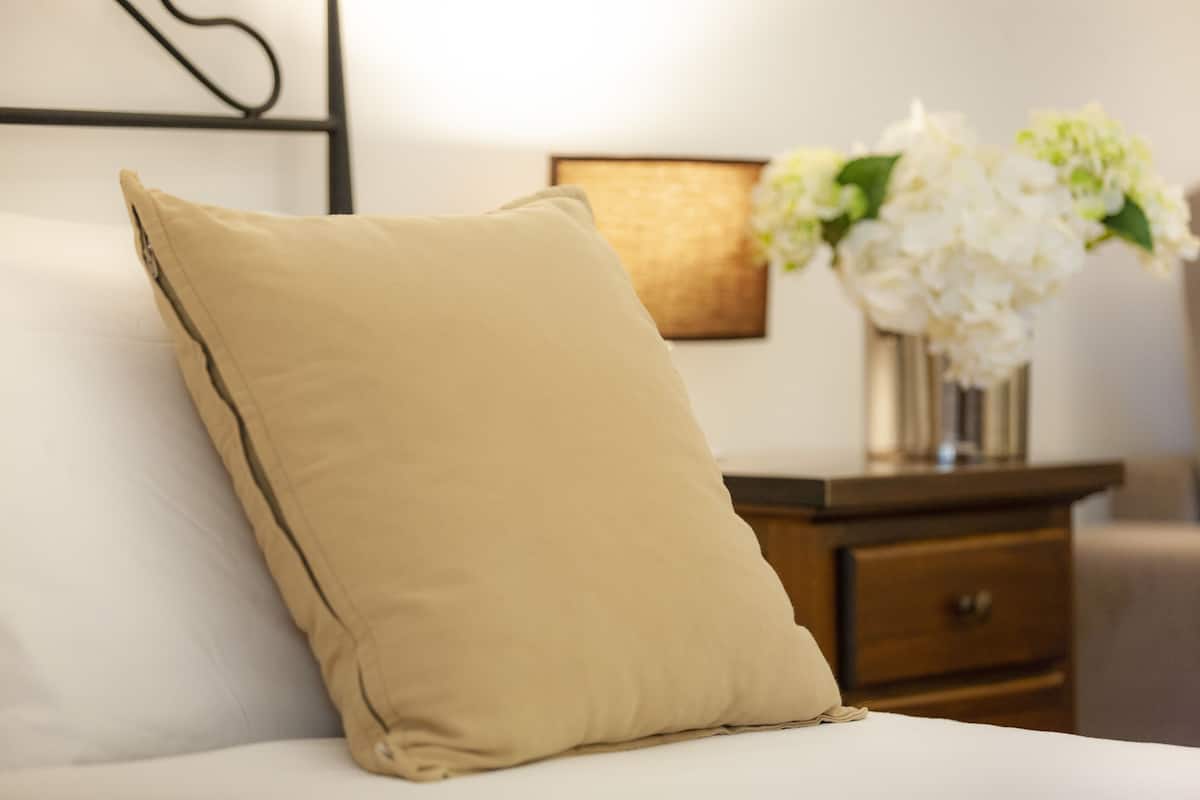 A bed with a beige pillow and a white mattress, a wooden nightstand with a vase of white flowers, and a framed picture on the wall.