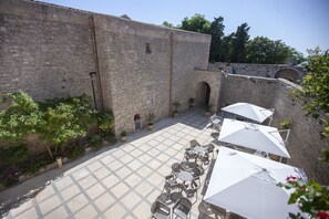 An outdoor patio with stone tables and white umbrellas, surrounded by stone walls and greenery.
